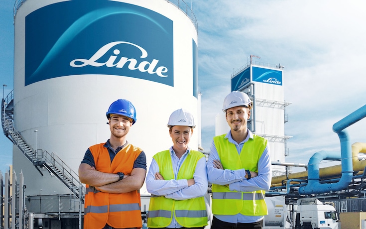 Drei Ingenieure mit Schutzhelmen und Warnwesten stehen vor einem Kraftwerk, mit einem großen Wasserstofftank der Marke Linde im Hintergrund.