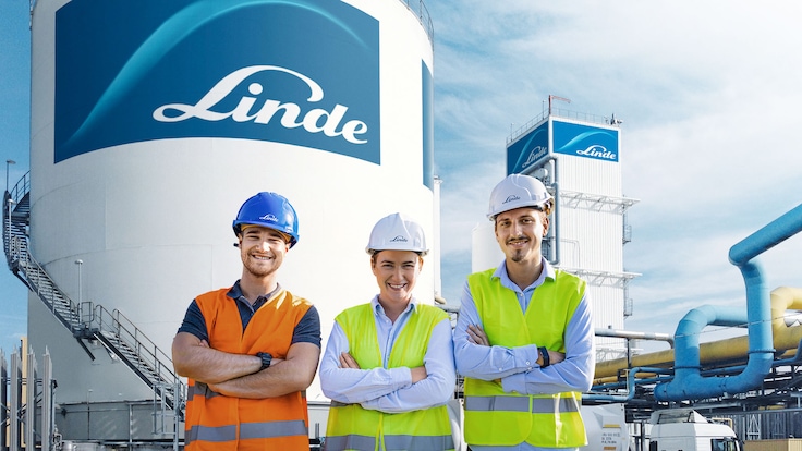 Drei Ingenieure mit Schutzhelmen und Warnwesten stehen vor einem Kraftwerk, mit einem großen Tank der Marke Linde im Hintergrund.