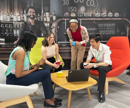 Gruppe von vier Absolventen in einer lockeren Geschäftsumgebung, die auf einen Laptop auf einem Couchtisch schauen.