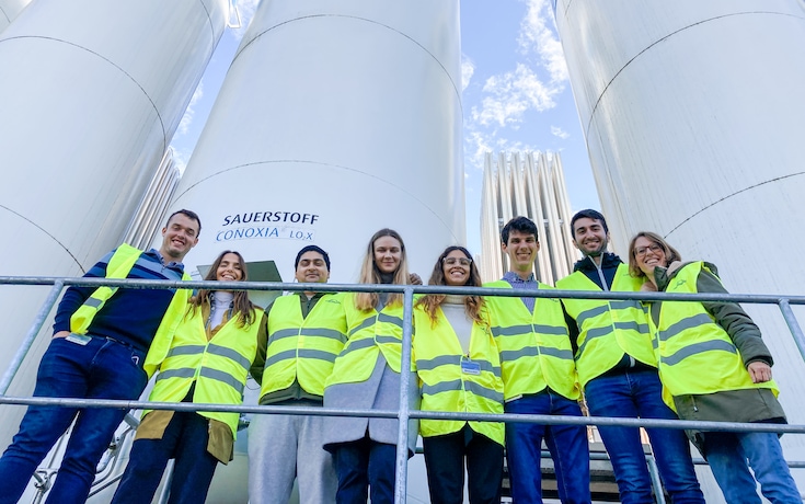 Eine Gruppe von acht jungen Absolventen in Warnwesten, die der Kamera zugewandt sind, mit hohen Gastanks der Marke Linde im Hintergrund.