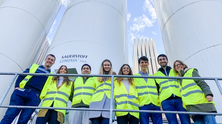 A group of eight young graduates in high visibility vests facing the camera with tall Linde branded gas cylinders in the background.