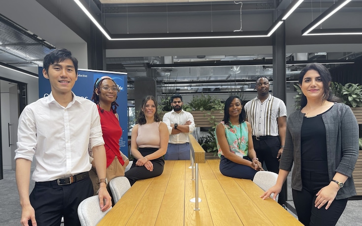 Eine vielfältige Gruppe von sieben jungen Absolventen versammelt um einen großen Tisch in einem Besprechungsraum.