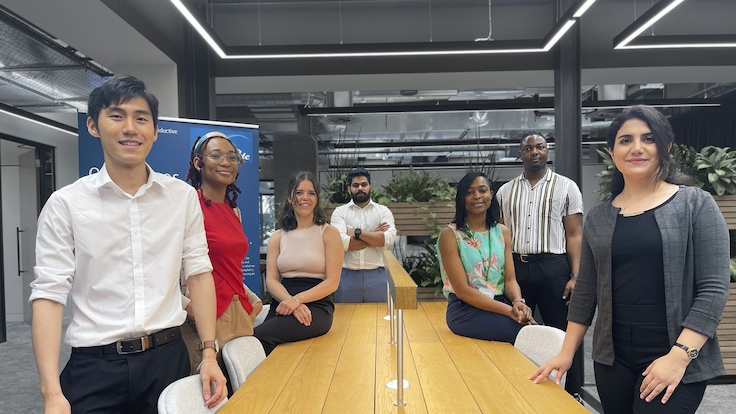 A diverse group of seven young graduates surrounding a large desk in a business meeting room.
