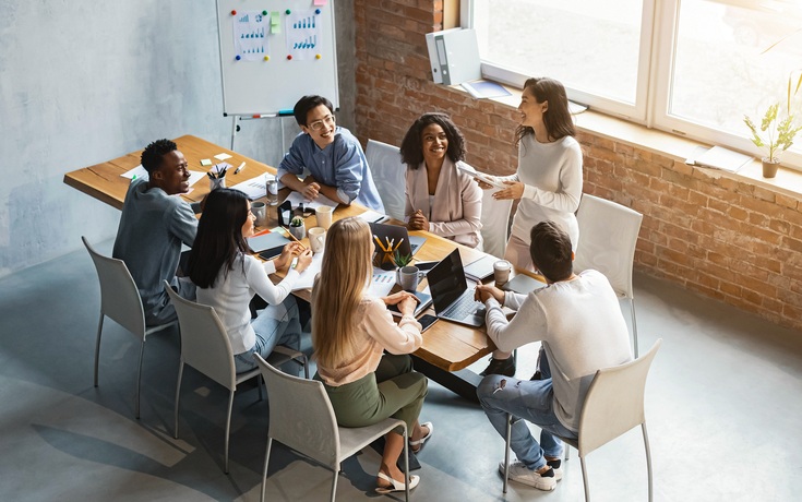 Eine Frau steht und teilt ihre Ideen mit Kollegen, die um einen Tisch in einem modernen Büro sitzen.