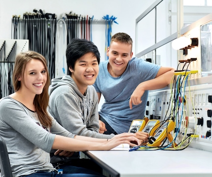 A group of young apprentices smile at the camera while working at an electronics workbench.