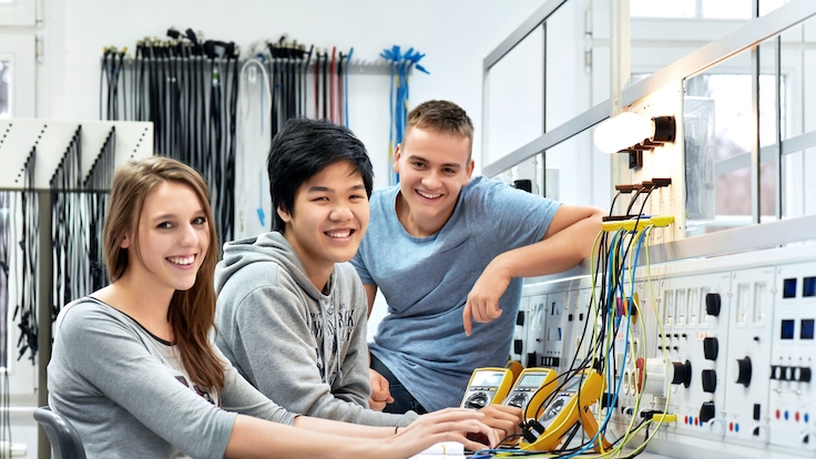Eine Gruppe junger Auszubildender lächelt in die Kamera, während sie an einer Elektronik-Arbeitsbank arbeitet