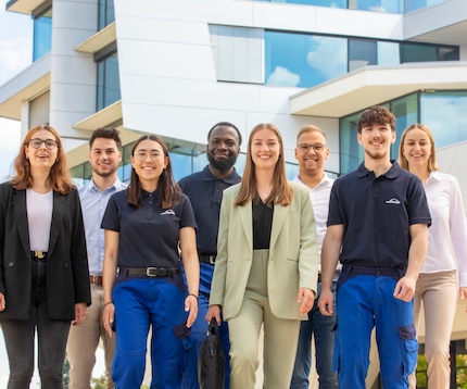 Eine Gruppe junger, lässig gekleideter Studenten lächelt in Gruppenformation vor einem der Gebäude von Linde in die Kamera.