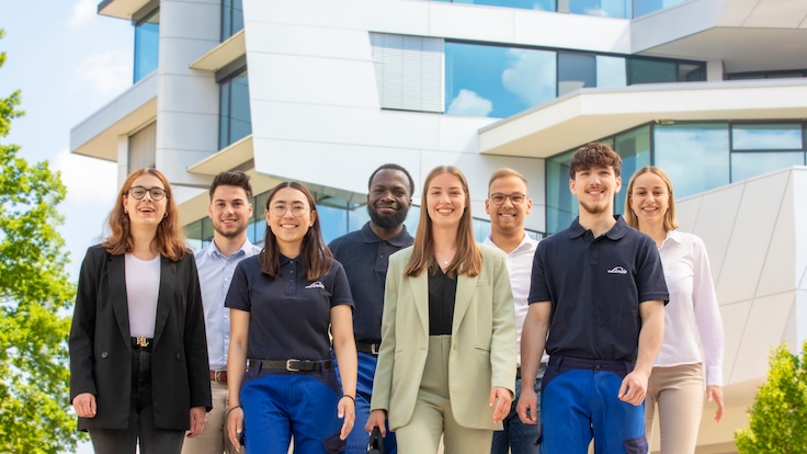 A group of casual, young students smile at the camera in group formation outside one of Linde’s buildings.