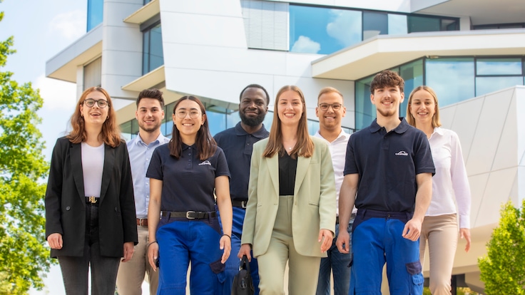 Eine Gruppe junger, lässig gekleideter Studenten lächelt in Gruppenformation vor einem der Gebäude von Linde in die Kamera.