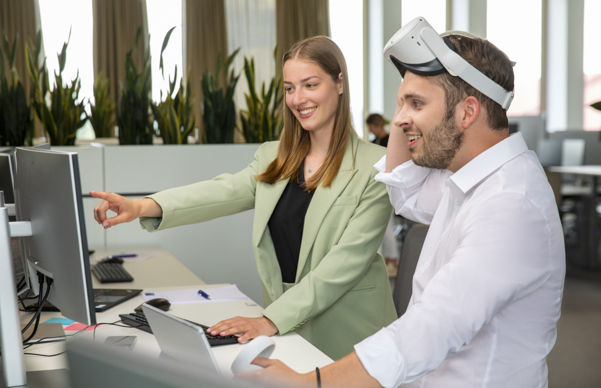 Two employees test a virtual reality system. One is wearing the VR headset and holds two handheld controls. The other looks at a monitor.