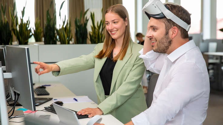 Two employees test a virtual reality system. One is wearing the VR headset and holds two handheld controls. The other looks at a monitor.