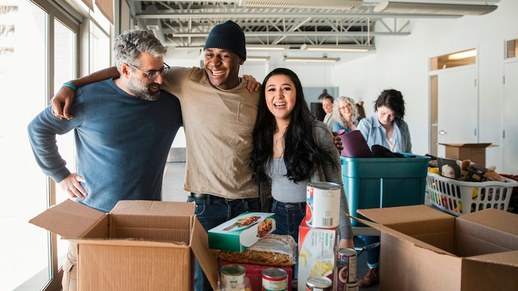 Drei Personen vor mehreren leeren Kartons und Lebensmitteln auf einem Tische employees in blue t-shirts at a corporate day of caring event holding signs."